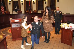 Newly promoted Col. Nick Saucier kneels for his daughter Livi, at left, and wife Bonny, at right, to apply the eagle rank insignia to his uniform during his Promotion and Investiture Ceremony on March 1 in Jackson. Maj. Gen. Janson Boyles, Adjutant General of the Mississippi National Guard, is in background at right.