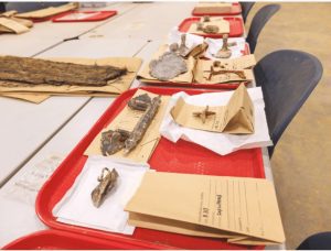 Objects found in Asylum Hill graves include a cap lifter (left, foreground) shaped like a calla lily and used to lift or replace a wooden panel that covered a glass viewing pane set in the coffin lid; and, beside it, a coffin handle. To the right of the coffin handle is a thumbscrew and escutcheon set; these were used to hold the coffin lid closed, as opposed to nailing it down.