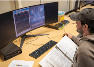 Syncom Space Services software test engineer Nicholas Payton Karno monitors a lab console at NASA’s Stennis Space Center displaying video footage of an RS-25 engine gimbal test, alongside data acquisition screens showing lab measurements.(Photo Credit: NASA/Danny Nowlin)