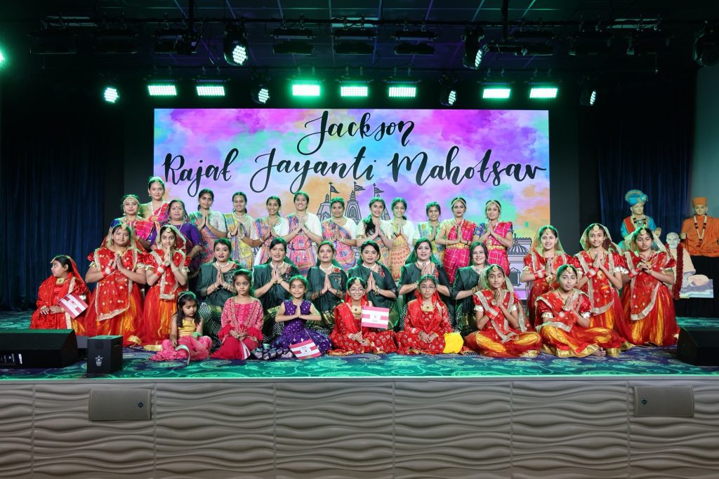 A stage photo of a large group of women and girls dressed in colorful traditional attire, posing together in front of a backdrop that reads “Jackson Rajat Jayanti Mahotsav.” This image shows cultural richness, youth involvement, and unity.