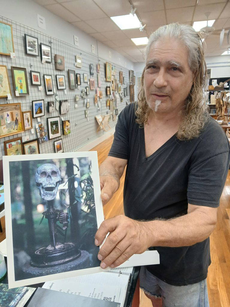 Armand Saiia displays a photograph of his sculpture “Jack the Humorist,” which incorporates a Model T Ford car jack, wheel hub, stone base, and a plaster skull.Photo by Roscoe Barnes III