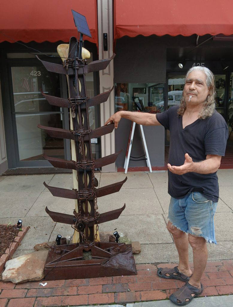 Sculptor Armand Saiia, ArtsNatchez’s Artist of the Month for June, poses with his large sculpture “Subjugation” in front of the gallery. The mixed-media work features wood carved to resemble bone, forged metal, chains, and solar-powered lighting.Photo by Roscoe Barnes III