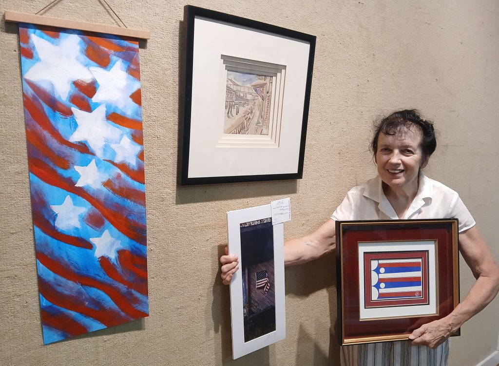 ArtsNatchez Gallery artist Jennifer Latetia White showcases patriotic-themed artwork ahead of Saturday's exhibit. The large banner on the left is by Carolyn Weir, while the framed piece on the wall is by Bradley Bruce. White holds a photograph by Leon Hollins (left) and a mixed-media piece by Bradley Bruce.