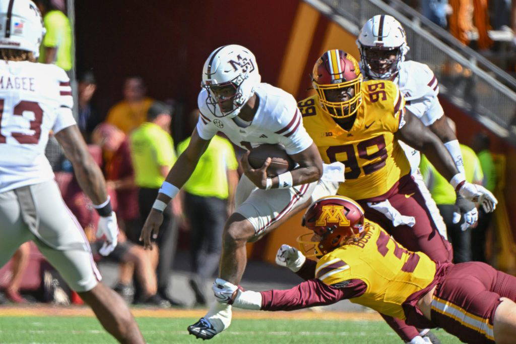 FILE - Mississippi State quarterback Kamario Taylor, center, leaps over Minnesota defensive lineman TJ Bush Jr. (3) in the first half during an NCAA college football game in Minneapolis, Saturday, Sept. 12, 2026. (AP Photo/Craig Lassig, File)